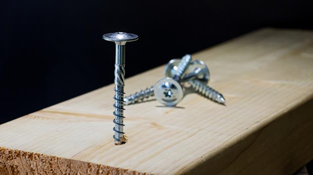 Close-up van schroeven op een houten plank, met één schroef deels ingeschroefd en meerdere schroeven liggend op de achtergrond tegen een zwarte achtergrond.