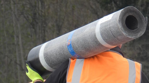 Bouwvakker in oranje veiligheidsvest draagt een grote rol dakbedekking op zijn schouder.