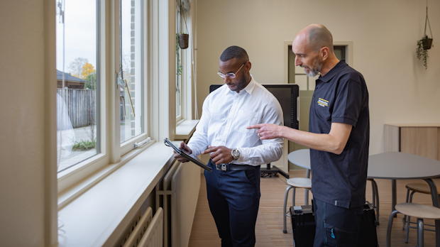 Technisch adviseur en opdrachtgever bekijken een tablet bij ramen in een utiliteitsgebouw.