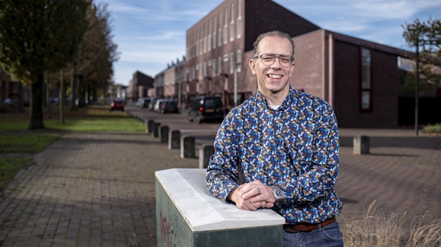 Man with glasses in a blue floral shirt leans on a concrete barrier on a brick street, with brick buildings and trees behind him.