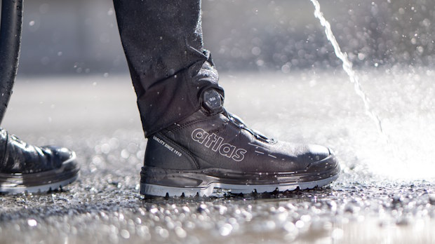 Close-up of a black Atlas boot stepping through a spray of water on a wet pavement.