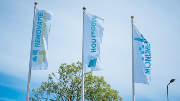 Three tall vertical flags fluttering in the wind against a clear blue sky, with light blue text and logos and a tree in the background.