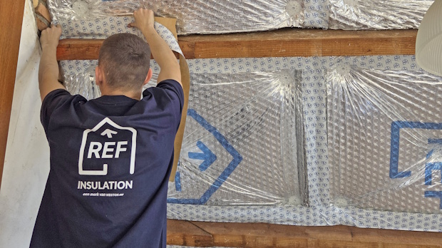 Worker wearing a REF Insulation shirt shows his back while attaching a wooden batten to a wall lined with reflective insulation panels in an attic.