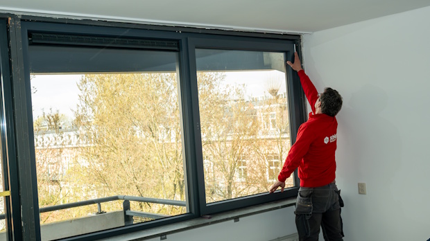 Worker in a red jacket installs or inspects a large window along a wall in a bright room.