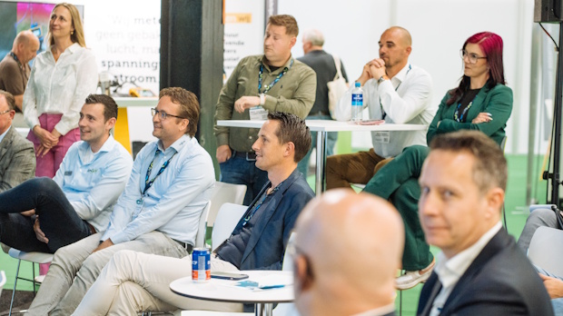 Group of professionals at a conference or workshop, seated around white tables and listening attentively to a speaker in the background.