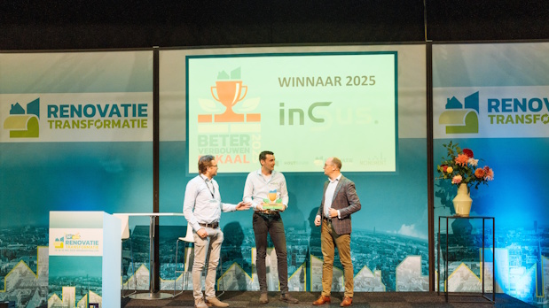 Three men on a stage at an award ceremony; one hands a trophy to another while a third watches, with Renovatie Transformatie banners on either side and a large screen displaying a trophy icon.