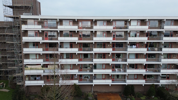 Front view of a multi-story brick apartment building with white balconies and red-framed doors; scaffolding on the left and leafless trees in front.