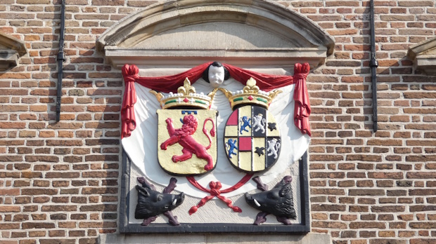Heraldic coat of arms mounted on a brick wall, featuring two shields with red lion and quartered designs, framed by red drapery.