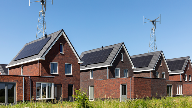Row of red brick houses with solar panels on the roofs and tall wind turbine masts against a clear blue sky, with grassy foreground.
