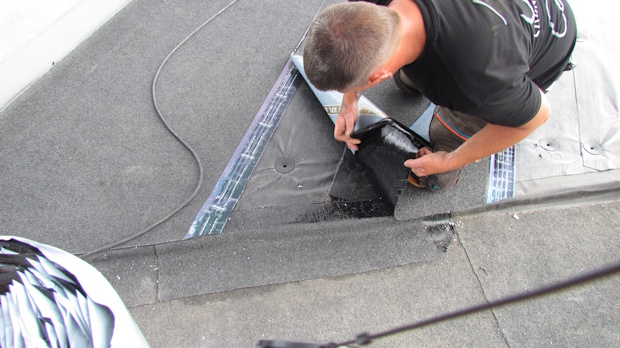 Roofer kneeling on a flat roof, sealing around a vent with a caulking gun.