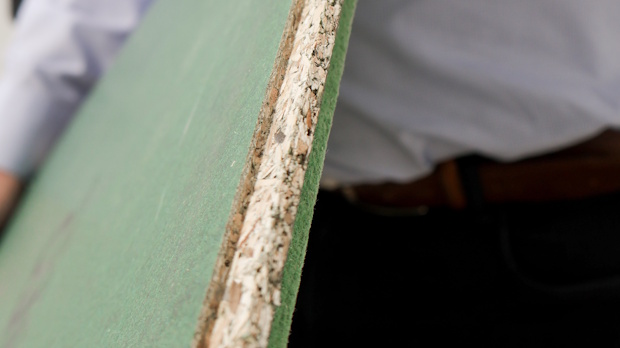 Close-up of a green board edge with exposed gypsum/insulation, held by a person in the background, indicating construction material.