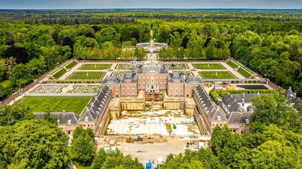 Aerial view of a grand historic chateau with symmetrical wings, formal terraces, and a green park, surrounded by trees.