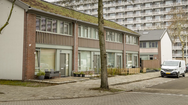 Row of brick two-story townhouses with front doors and small gardens, a tree in the foreground, and a white van parked on the street; a tall apartment block looms in the background.