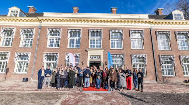 Group of people posing for a photo on a red carpet in front of a large brick building on a sunny day, likely a ceremony or event.