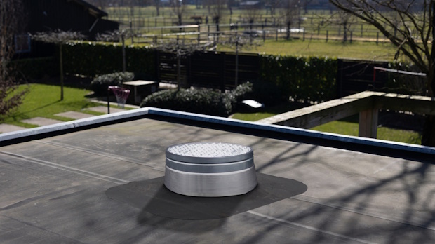 Circular metal roof vent on a flat rooftop with a shadow, background shows a fenced grassy yard and trees.