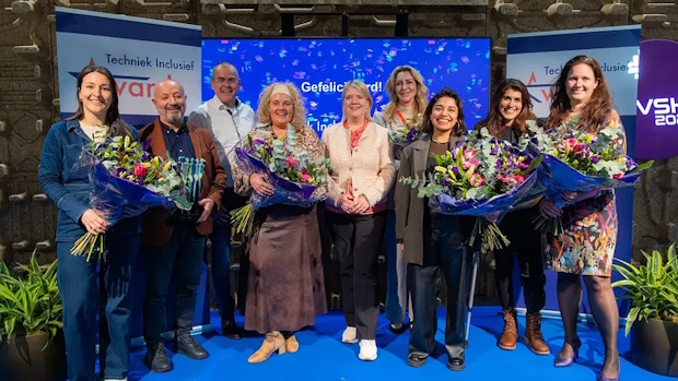 Eight people on a blue stage, holding bouquets and smiling, with 'Techniek Inclusief' banners in the background.