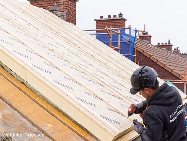 Construction worker on a sloped roof, drilling into insulation panels with scaffolding and brick houses in the background.