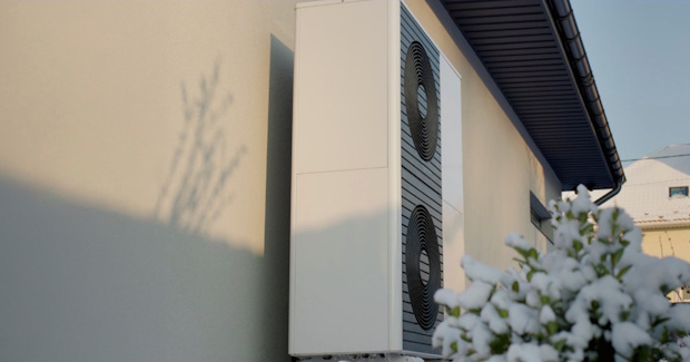 Outdoor HVAC condenser unit mounted on the side of a beige building, with snow-covered bushes in the foreground.