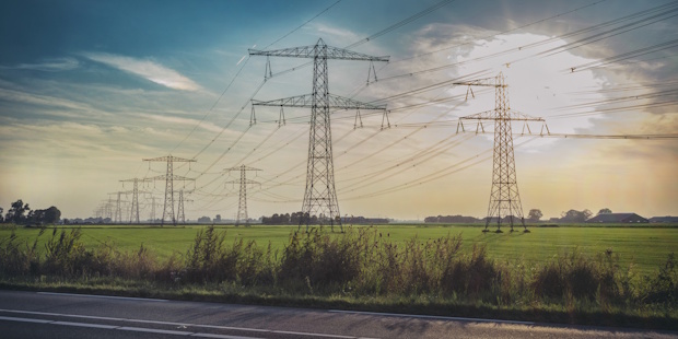 High-voltage transmission towers stretch across a flat rural field near a road at sunset.
